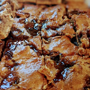 May include: A tray of baked brown square bars with a dense, textured surface and visible dark, glossy, jam-like filling between the layers. The baked goods have a golden-brown color with irregular edges and a slightly cracked, crumbly top texture.