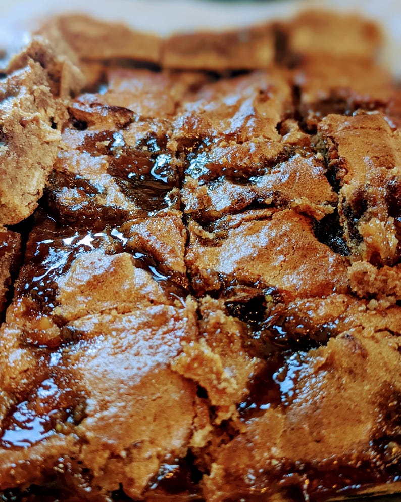 May include: A tray of baked brown square bars with a dense, textured surface and visible dark, glossy, jam-like filling between the layers. The baked goods have a golden-brown color with irregular edges and a slightly cracked, crumbly top texture.
