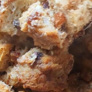 May include: A close-up view of a golden-brown bread pudding featuring chunks of toasted bread and dark raisins. The texture is soft and porous, with visible variations in color ranging from light tan to deep brown throughout the baked mixture.