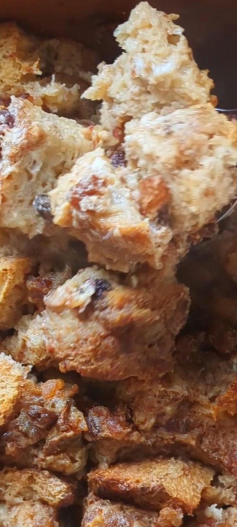 May include: A close-up view of a golden-brown bread pudding featuring chunks of toasted bread and dark raisins. The texture is soft and porous, with visible variations in color ranging from light tan to deep brown throughout the baked mixture.