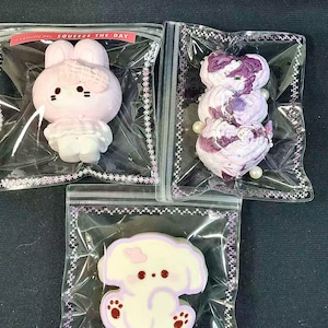 May include: Three individually packaged, decorative items. The top left item is a pink bunny with a bow. The top right item is a stack of purple and white floral shapes. The bottom item is a white dog with purple accents. Each item is in a clear plastic bag.