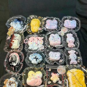 May include: A collection of decorative soaps in various shapes, including flowers, bears, and bunnies, each encased in clear plastic with decorative edges. Colors range from pastel pinks and blues to vibrant yellows and browns. The soaps are arranged on a black surface.