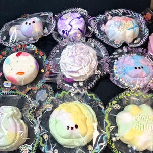 May include: A collection of nine round, individually wrapped decorative charms featuring pastel colors, marbled patterns, and small character faces. Each charm is encased in clear plastic with a decorative scalloped border and small printed icons.