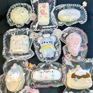 May include: A collection of colorful, plush toys in clear plastic packaging. The toys include a mouse, ice cream cone, cloud, and cake, all with cute, cartoonish designs. Each toy is individually packaged with decorative trim and charms.