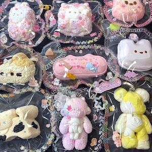 May include: A collection of various squishy toys in individual clear plastic packaging. The items include animal-shaped figures, a pink rectangular case, and a yellow bear-like character, all featuring pastel colors, textured surfaces, and small decorative stickers.