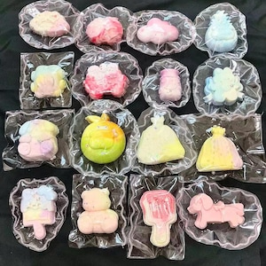 May include: An assortment of colorful, shaped bath bombs in clear plastic packaging. The collection includes cloud, flower, and animal shapes in pastel pink, yellow, and blue hues. The bath bombs are arranged on a dark surface.