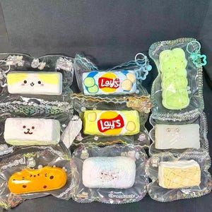 May include: A collection of colorful, squishy toys in clear plastic packaging. The toys are shaped like food items, including a yellow rectangle with the Lay's logo, a green stack of fries, and other cute designs.