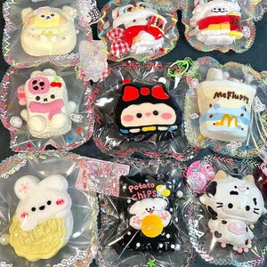 May include: A collection of colorful, cartoon-style plush toys in clear plastic packaging. The toys include a white bear, a red and white lion, a pink and white cat, a black and white character, and a McFlurry cup.