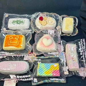 May include: An assortment of colorful, packaged, decorative food items. The items include a white rectangular item with green sprinkles, a round item with a cherry, and a rectangular item with the text "Sweet".