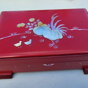 May include: A red wooden box with a hinged lid decorated with a rooster and two chicks made of mother-of-pearl inlay. The box has a butterfly design on the front and sits on four small feet.