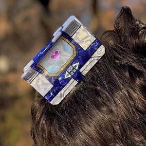 May include: A rectangular hair clip featuring a blue and white geometric pattern with a central screen display showing a pink heart icon. The accessory is secured in dark brown hair, showcasing a metallic finish and a modern, angular design.