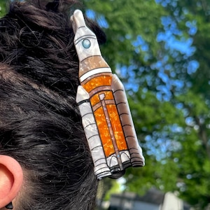 以下が含まれることがあります： スペースロケットの形をしたヘアクリップ。白と真珠のような光沢のあるボディに、鮮やかなオレンジ色の中央セクション、上部近くの小さな青い円形のディテール、そしてロケット構造の基部と側面に沿った黒いアクセントが特徴です。