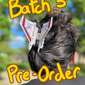 May include: A hair claw clip shaped like a white and grey marbled spaceship with red wing accents and purple heart details. The text "Batch 3" appears at the top and "Pre-Order" at the bottom in yellow font with a brown outline.