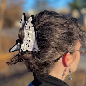May include: A space shuttle-shaped hair clip in black and white, worn in the hair. The clip is detailed with lines and shapes, and is set against a blurred outdoor background.
