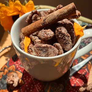 May include: A white mug filled with candied pecans and a cinnamon stick. The pecans are coated in a light brown glaze. The mug sits on a patterned cloth with a sunflower and orange flower petals.