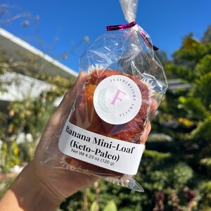 May include: A banana mini-loaf in a clear cellophane bag, tied with a pink ribbon. The label reads "Banana Mini-Loaf (Keto-Paleo)" and "Net Wt 120 g". The logo features a green wreath and the letter "F".