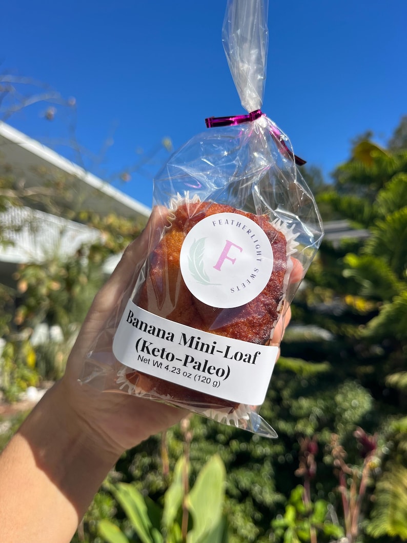 May include: A banana mini-loaf in a clear cellophane bag, tied with a pink ribbon. The label reads "Banana Mini-Loaf (Keto-Paleo)" and "Net Wt 120 g". The logo features a green wreath and the letter "F".