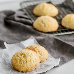 May include: Close-up of two light brown, round biscuits on a white piece of parchment paper. The biscuits are slightly browned and have a soft, crumbly texture.