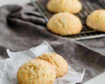 May include: Close-up of two light brown, round cookies on a white parchment paper. The cookies are slightly browned and have a soft, crumbly texture.