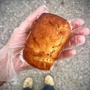 May include: A freshly baked, golden-brown rectangular muffin in a white paper case. The muffin is held in a gloved hand. The image is taken from above, with the person's feet visible in the background. The muffin has a textured surface.