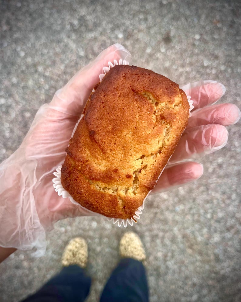 May include: A freshly baked, golden-brown rectangular muffin in a white paper case. The muffin is held in a gloved hand. The image is taken from above, with the person's feet visible in the background. The muffin has a textured surface.