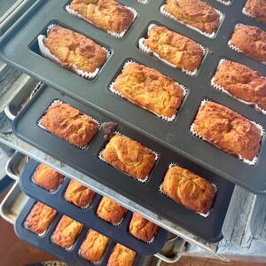 May include: A close-up of several baking trays filled with golden-brown mini loaves of bread. Each loaf is baked in a white paper case. The trays are stacked, indicating a fresh batch of baked goods, possibly for sale.