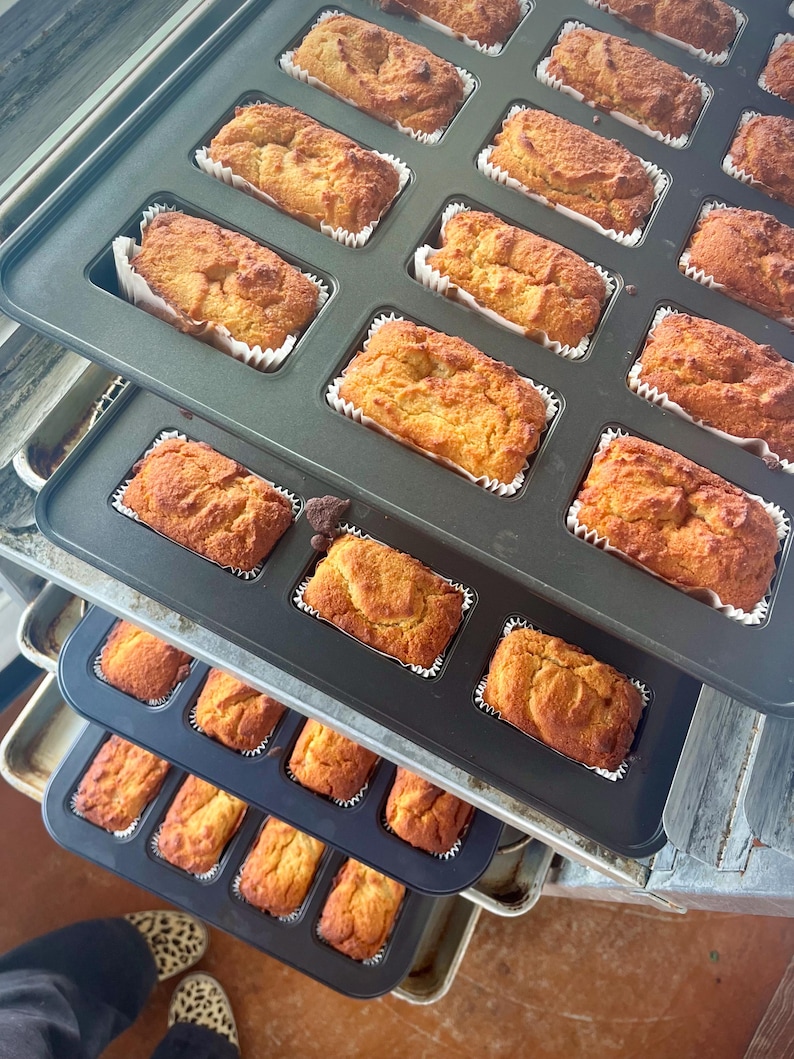 May include: A close-up of several baking trays filled with golden-brown mini loaves of bread. Each loaf is baked in a white paper case. The trays are stacked, indicating a fresh batch of baked goods, possibly for sale.