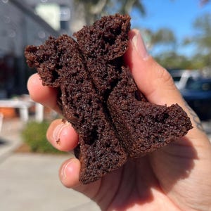May include: A close-up of a chocolate brownie broken in half, showing the inside of the brownie. The brownie is dark brown and has a slightly crumbly texture.