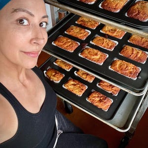 May include: Several trays of freshly baked mini loaves of bread, each in a white paper case, are arranged on a metal rack. The loaves are golden brown. The image suggests a home bakery or kitchen setting, with the loaves ready for sale.