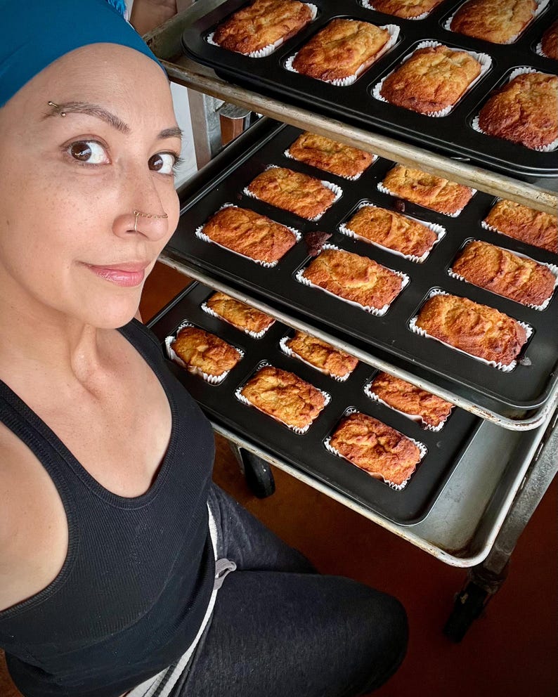 May include: Several trays of freshly baked mini loaves of bread, each in a white paper case, are arranged on a metal rack. The loaves are golden brown. The image suggests a home bakery or kitchen setting, with the loaves ready for sale.