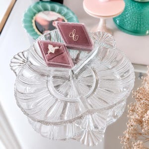 May include: A clear glass divided serving dish with a scalloped edge design. Two diamond-shaped mauve velvet ring boxes are placed inside the dish, with one box holding a silver-toned ring featuring a pear-shaped stone and a gold-toned letter C.