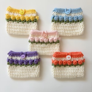Puede incluir: Cinco bolsos de ganchillo hechos a mano en varios colores: amarillo, azul, rosa, morado y naranja. Cada bolso presenta un cuerpo color crema con una fila de flores texturizadas coloridas y un cierre de botón a juego. Los bolsos miden aproximadamente 10 cm de ancho y 13 cm de alto.