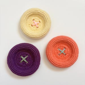 Puede incluir: Tres posavasos de ganchillo hechos a mano con diseño de botón. Los posavasos son en tonos amarillo, morado y naranja, cada uno con un centro de punto de cruz contrastante. El posavasos amarillo tiene un punto de cruz rosa, el morado uno verde.