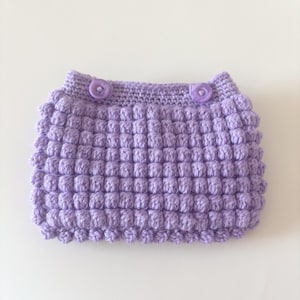 May include: A light purple crochet garment featuring a textured bobble stitch pattern throughout the main body. The waistband is constructed with a flat stitch and includes two matching light purple circular buttons positioned at the front.