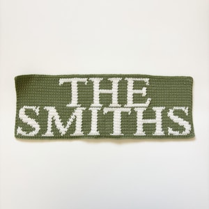 May include: Olive green crocheted banner with the text "THE SMITHS" in white. The banner is rectangular and appears to be handmade. The text is centered and clearly visible against the green background.