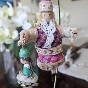 May include: A handmade ornament featuring two dolls dressed in festive attire. The larger doll is wearing a pink dress with gold trim and a white lace scarf. The smaller doll is wearing a green dress with a white lace trim. Both dolls have brown hair and are wearing hats.