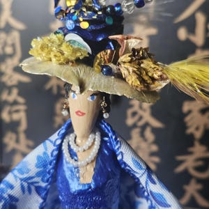 May include: A wooden doll with blue eyes and red lips wears a blue velvet dress and a large hat decorated with seashells, feathers, and beads.