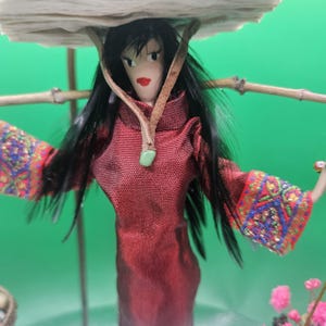 May include: A doll with long black hair and a red dress, carrying a load on its head. The doll's sleeves have colorful embroidered patterns. A green background provides a contrasting backdrop for the doll.