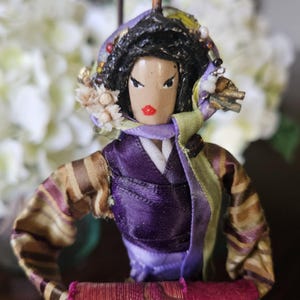 May include: A handmade doll with black hair, wearing a purple and brown kimono with a green and purple scarf. The doll has a painted face with red lips and black eyes.