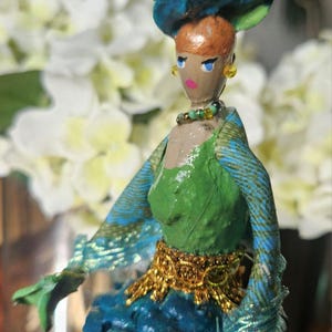 May include: A handmade doll with a green dress, blue and gold patterned shawl, and a blue hat with yellow flowers. The doll has a painted face with brown hair and blue eyes.