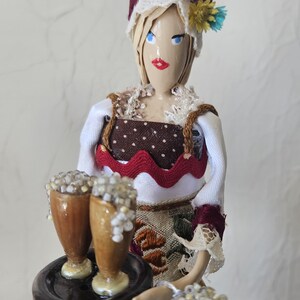 May include: A handcrafted doll dressed in traditional attire, holding a tray with two beer glasses. The doll has a painted face, a white lace-trimmed bonnet, and a brown and white polka-dot bodice. The beer glasses are filled with a foamy beverage.