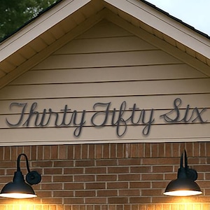May include: Metal house number sign in a cursive font that reads "Thirty Fifty Six" mounted on a beige house exterior. The sign is above a brick wall and a white garage door. Two black outdoor lights illuminate the area.
