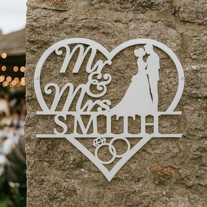 May include: White metal wedding sign in the shape of a heart, with the words "Mr & Mrs SMITH" and a silhouette of a bride and groom. Two rings are at the bottom of the heart. The sign is mounted on a stone wall.