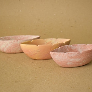 May include: Three oval-shaped stone bowls with a smooth, matte finish. The bowls are arranged in a row and feature varying shades of soft pink, peach, and light coral with subtle white marbling patterns throughout the natural stone material.