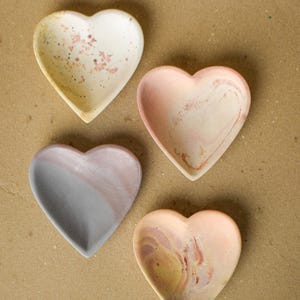 May include: Four heart-shaped trinket dishes arranged in a group. One dish is cream with pink speckles, one is a pale pink swirl, one is split between grey and soft pink, and the final dish features a marbled blend of peach, pink, and yellow tones.