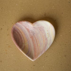May include: A heart-shaped dish featuring a smooth, polished surface with swirling patterns of cream, soft pink, muted purple, and earthy brown tones. The bowl has a concave center and a rounded rim, showcasing natural stone-like color variations.