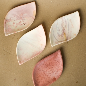 May include: Four leaf-shaped stone dishes arranged in a cluster. Each dish features a unique natural pattern with varying shades of pink, cream, and white. The surfaces show distinct mineral textures, swirls, and speckles characteristic of polished stone.