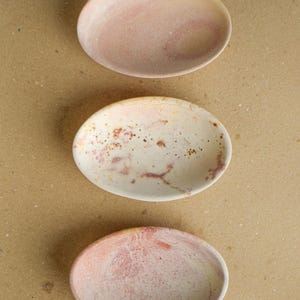 May include: Three oval-shaped dishes arranged vertically. The top dish is a solid pale pink. The middle dish is off-white with speckled rust-colored and pink markings. The bottom dish features a marbled blend of soft pink, peach, and cream tones.