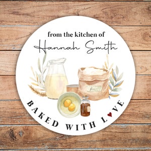 May include: Round white sticker with a watercolor illustration of baking ingredients, including flour, eggs, vanilla extract, and milk. The text reads "from the kitchen of Hannah Smith BAKED WITH LOVE".