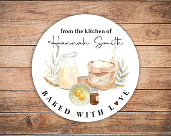 May include: Round white sticker with a watercolor illustration of baking ingredients, including flour, eggs, vanilla extract, and milk. The text reads "from the kitchen of Hannah Smith BAKED WITH LOVE".